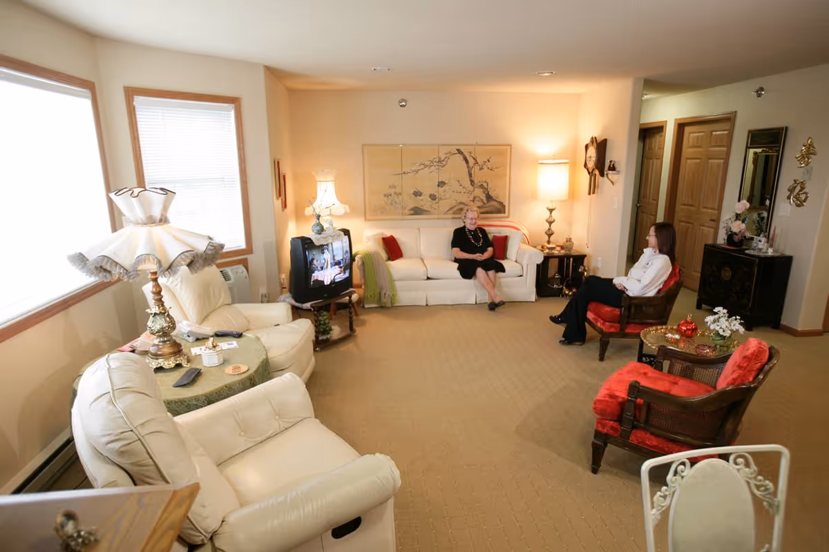 A bright, carpeted living room with sofas and armchairs, lamps, a television, and two women seated and talking.