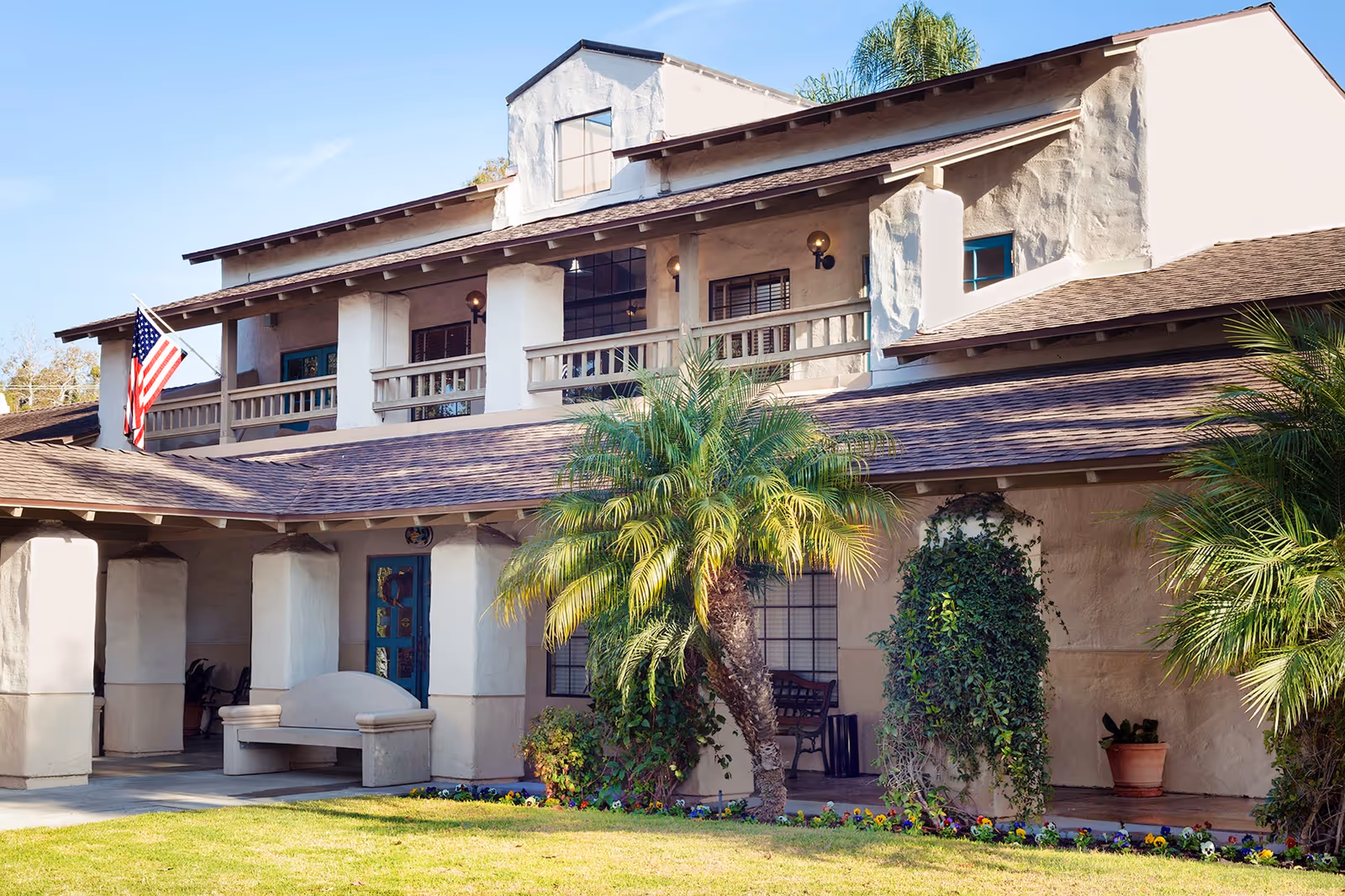 Exterior view of a two-story building with a covered porch, palm trees, and an American flag. The building has a beige stucco finish and a shingled roof, with a bench and potted plants near the entrance.