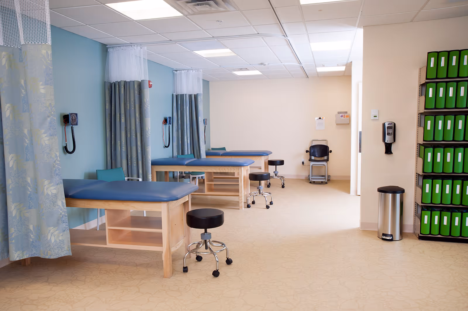 A clinical treatment room with three examination tables separated by blue and white privacy curtains. Each table has a black rolling stool and a green chair nearby. The room has light blue and beige walls, a beige patterned floor, and a wall-mounted blood pressure monitor. On the right side, there is a shelf filled with green binders, a hand sanitizer dispenser, and a silver trash can.