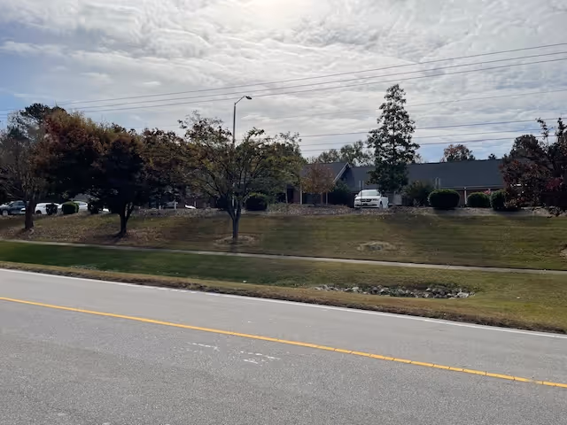 View of a single-story building partially obscured by trees and bushes, with a grassy area and a road in the foreground under a cloudy sky.