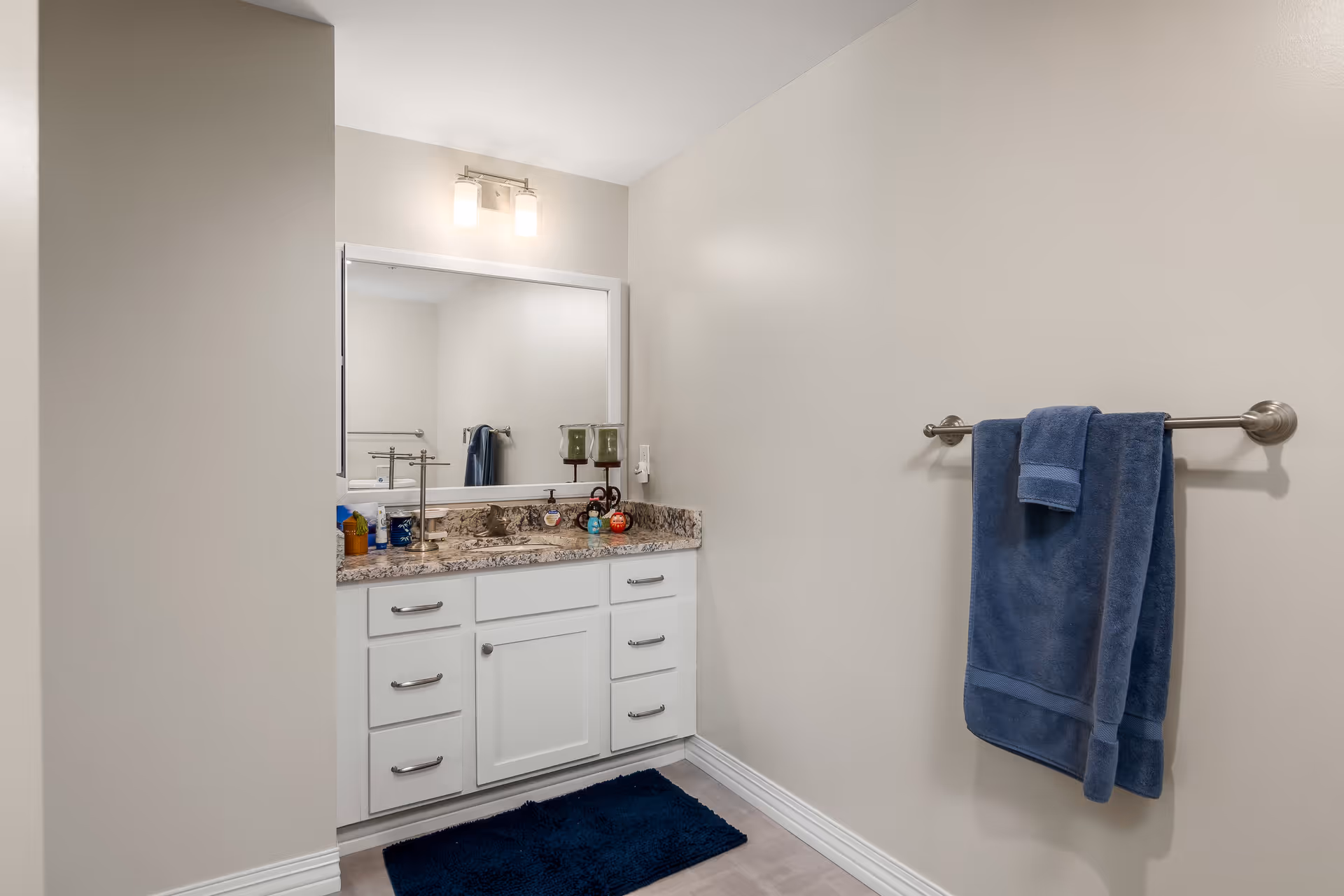 A clean bathroom vanity area with a large mirror above a granite countertop. The vanity has white cabinets and drawers with silver handles. On the countertop are various toiletries and two green candle holders. A silver towel rack on the adjacent wall holds two blue towels. The walls are painted light beige, and there is a dark blue bath mat on the floor.