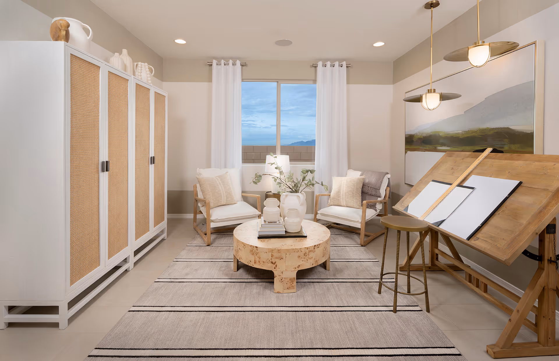 A bright and cozy interior room featuring two cushioned armchairs with pillows, a round wooden coffee table with decorative vases and books, a large white cabinet with woven panel doors, a drafting table with a stool, and a large landscape painting on the wall. The room has a window with white curtains showing a blue sky and mountains outside.
