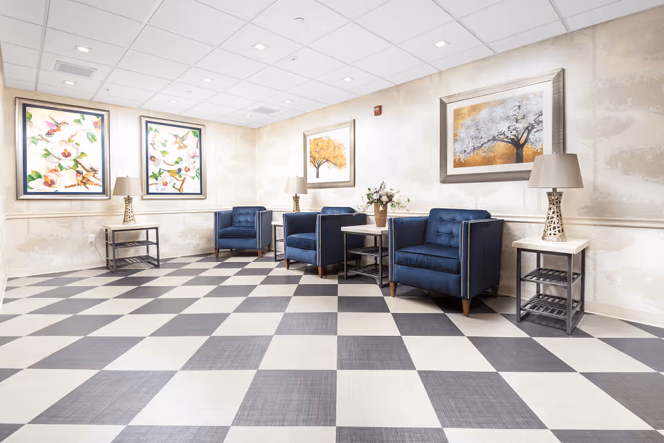Bright waiting room with blue armchairs, side tables, lamps and framed artwork on a checkered floor.