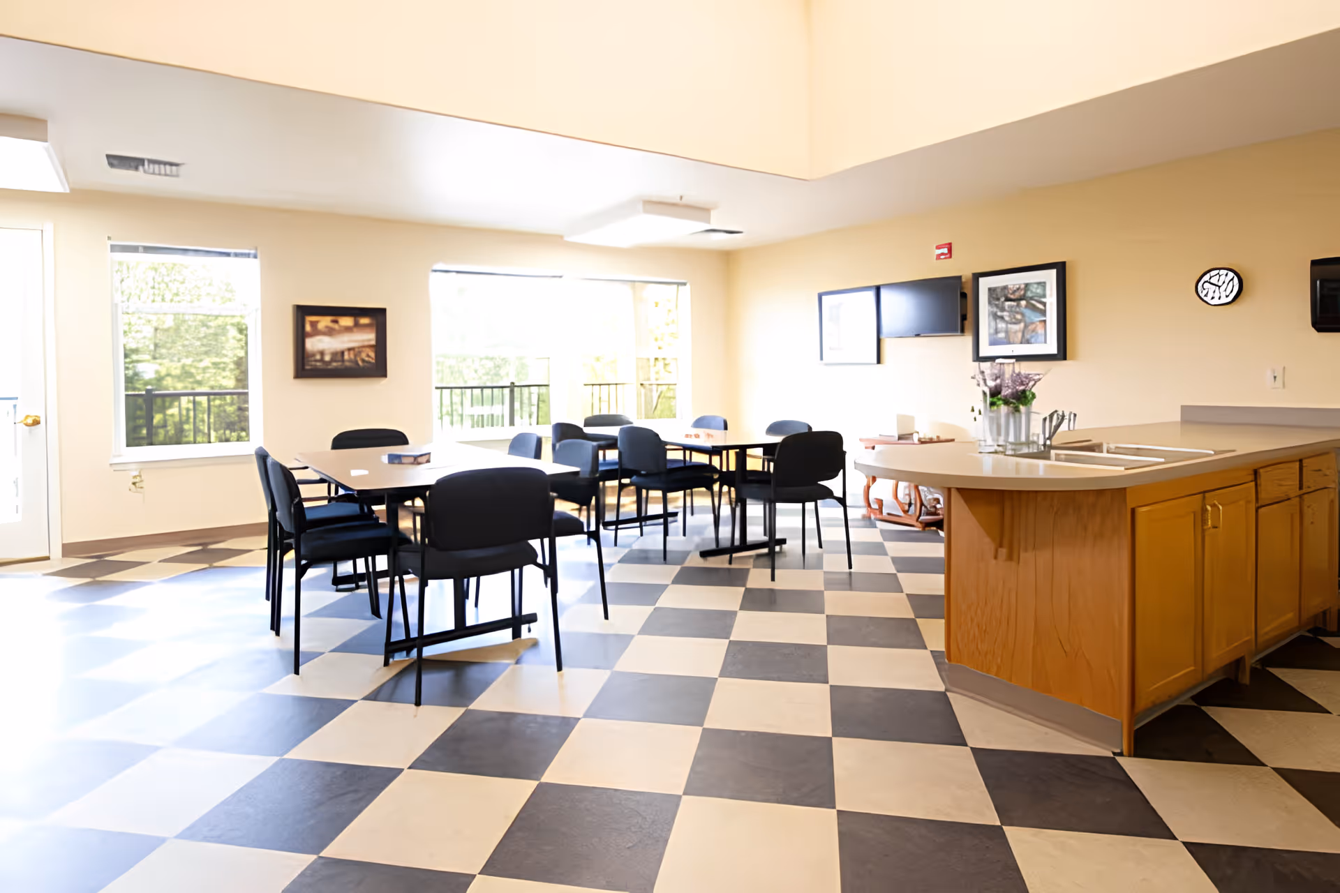 A bright room with large windows letting in natural light, featuring black and white checkered flooring, several tables with black chairs, a wooden counter with a sink and flowers in a vase, and framed artwork on the beige walls.