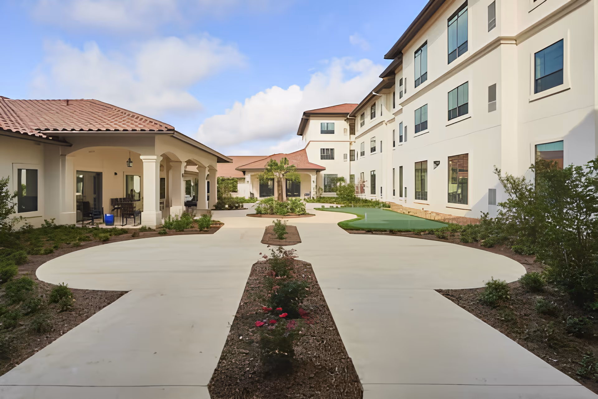 Outdoor courtyard area of a senior living facility with a wide concrete pathway, landscaped garden beds, and a putting green. The buildings surrounding the courtyard have multiple windows and a covered patio area with seating. The sky is partly cloudy.
