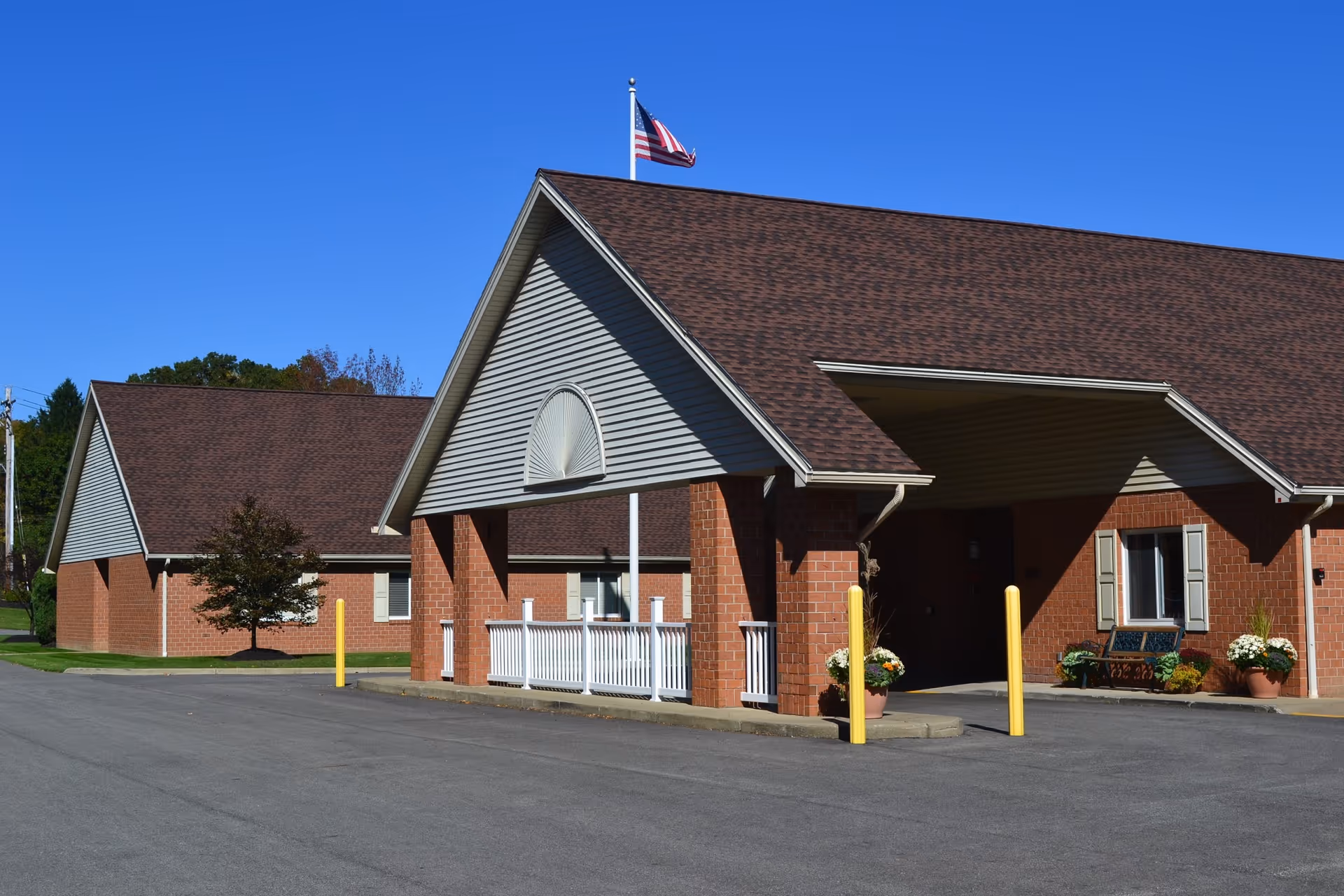 Brick single-story senior care building with a covered entrance, American flag, and potted plants under a clear blue sky.