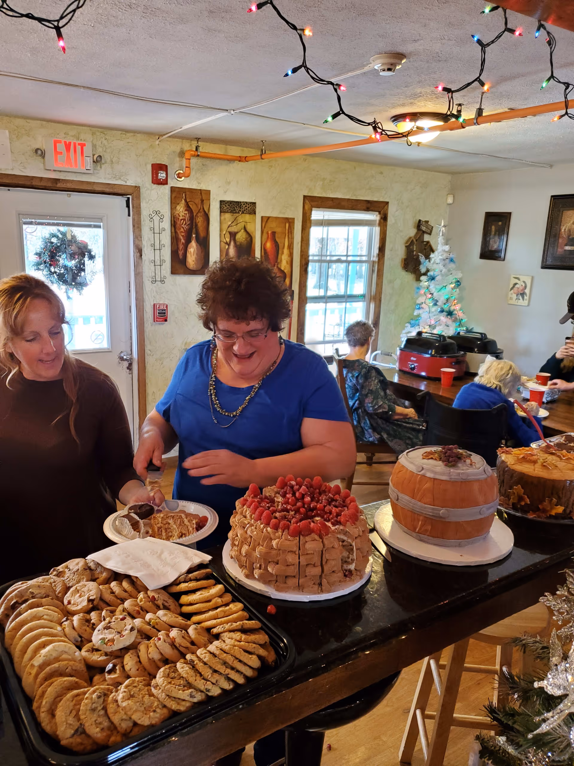 Two women standing at a counter with a variety of desserts including cookies and three decorated cakes. One woman is serving a slice of cake onto a plate. In the background, there are elderly people sitting at a table near a small decorated Christmas tree. The room has festive string lights hanging from the ceiling and wall decorations.