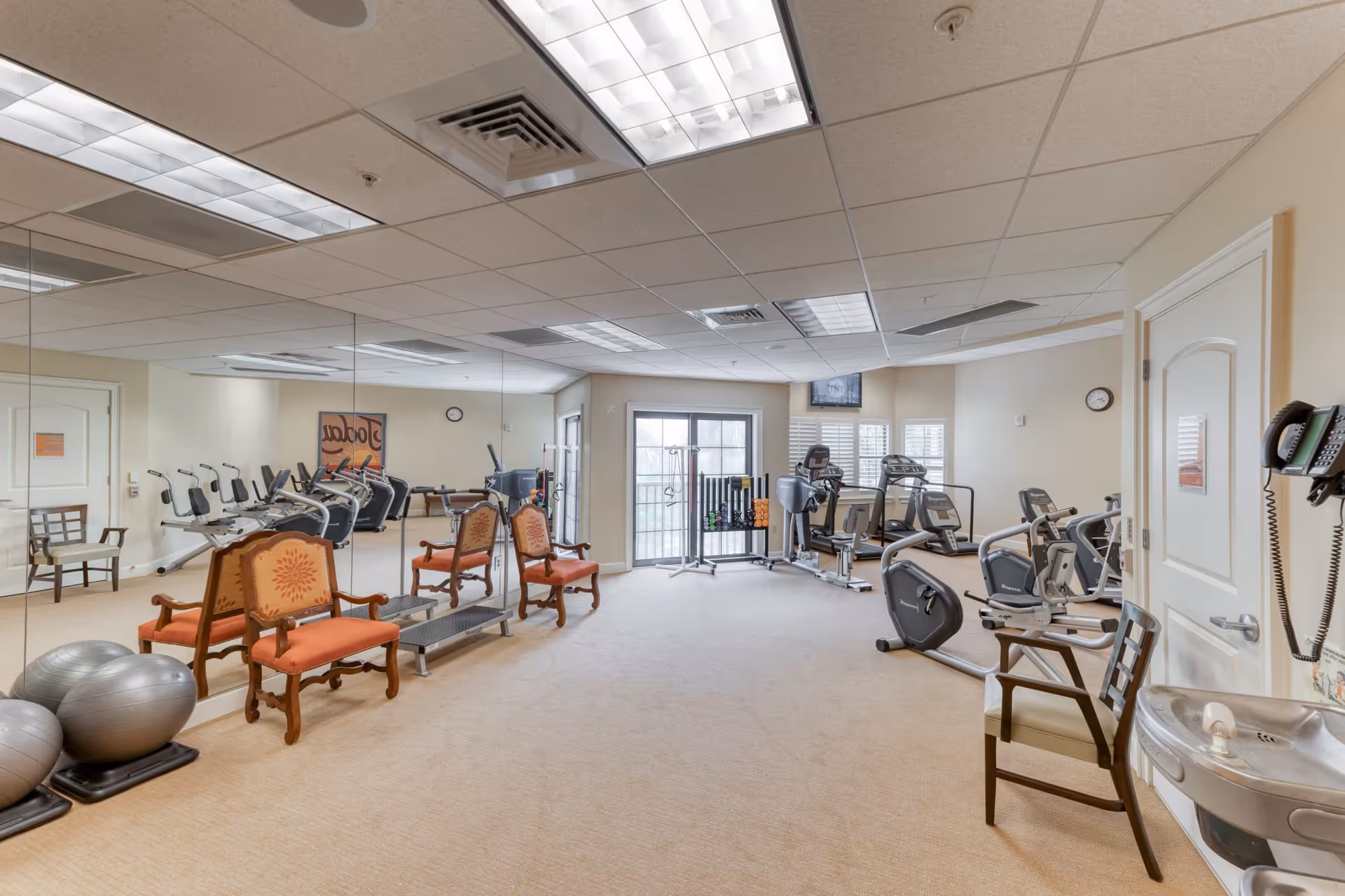 Spacious fitness room with cardio machines, exercise balls, mirrored wall and seating in a senior living facility.
