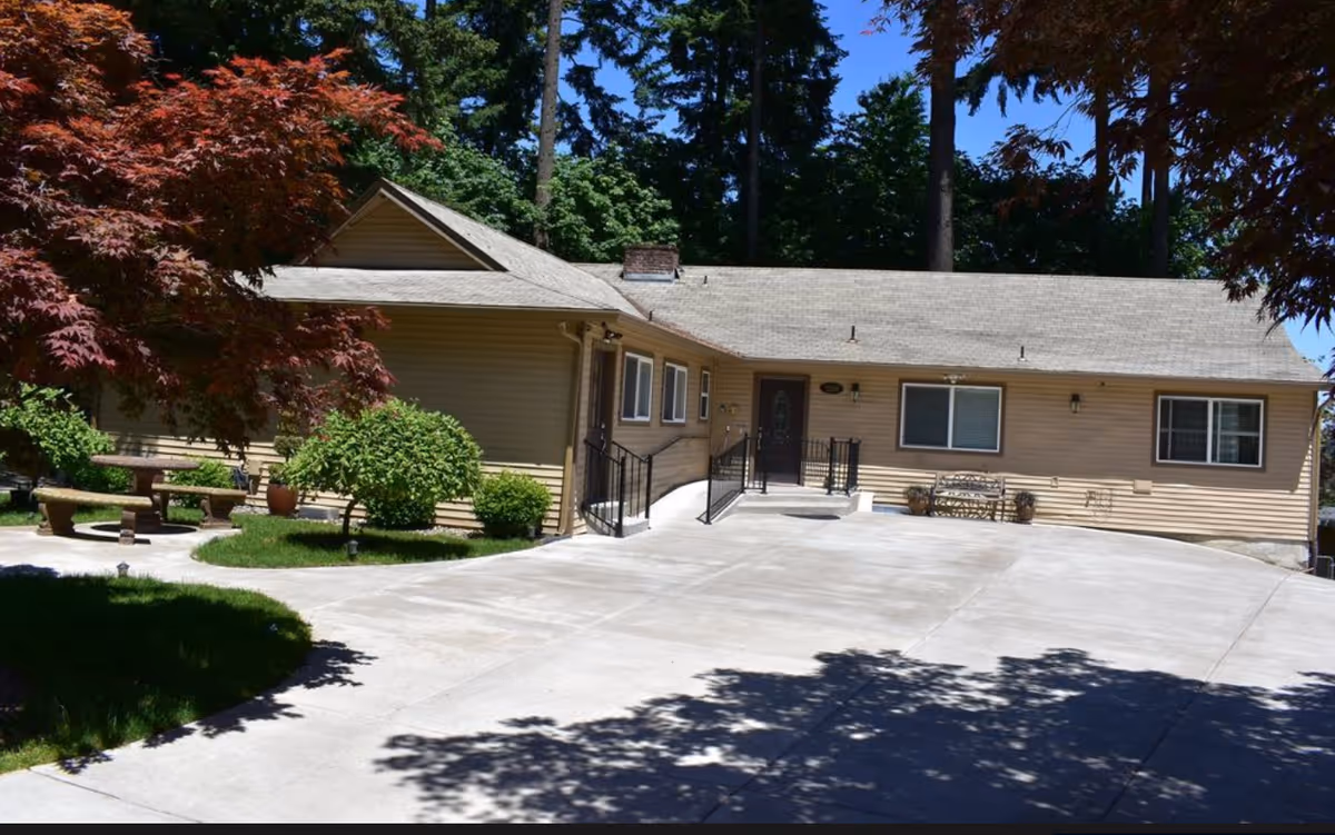 Single-story beige house front with a wide concrete driveway, entrance ramp and landscaped trees and shrubs.