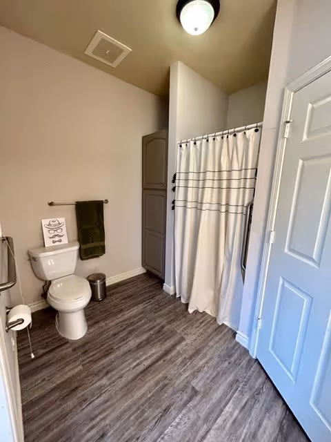 A clean bathroom with a white toilet, a green towel hanging on a towel rack, a small trash can, a shower with a white curtain featuring horizontal black stripes, and a closed white door. The floor has wood-like vinyl planks and the walls are painted beige.