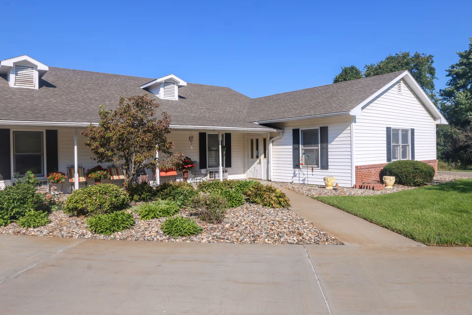 Exterior view of a single-story white building with a gray shingled roof, black shutters, and a small porch with chairs and potted plants. The front yard features a landscaped area with bushes, a small tree, and rocks, along with a concrete driveway and walkway leading to the entrance. The sky is clear and blue.