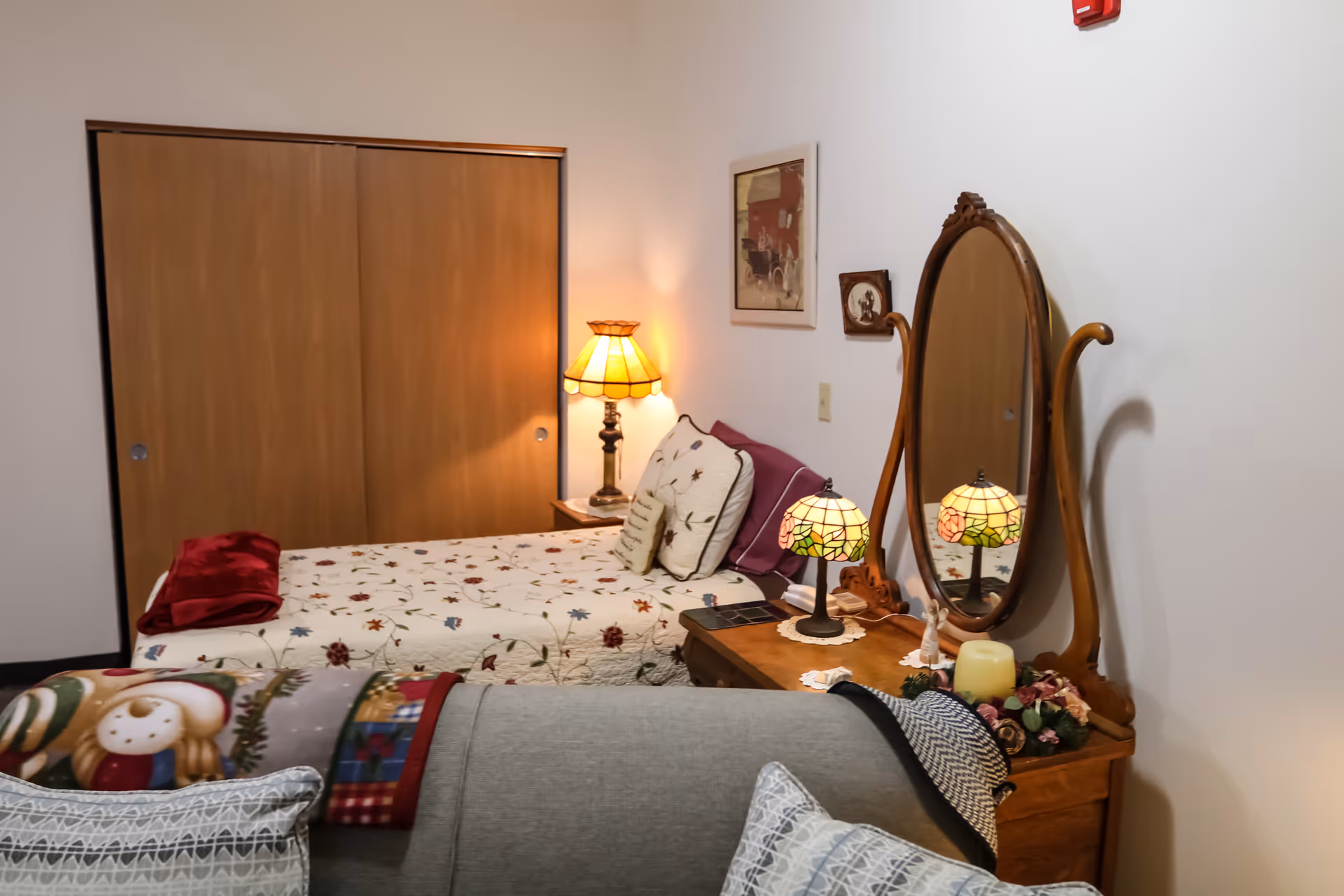 A cozy bedroom with a bed covered in a floral quilt and pillows, a wooden dresser with an oval mirror, two Tiffany-style lamps, a table lamp on a nightstand, and a couch with patterned pillows and a holiday-themed blanket in the foreground.