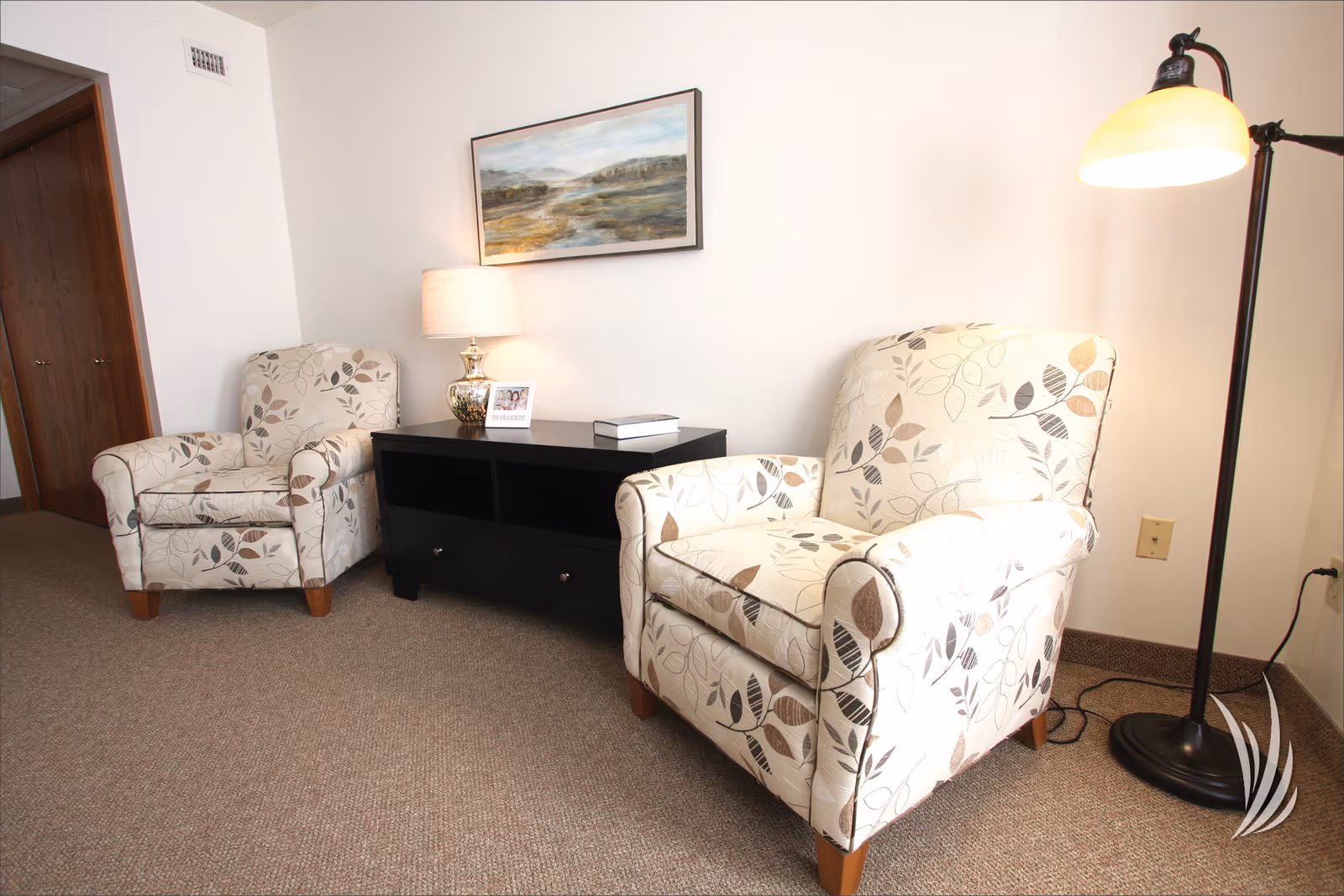 A cozy sitting area with two beige armchairs featuring a leaf pattern, a black side table with a lamp, a framed photo, and a book on it. A floor lamp with a yellow shade stands to the right, and a landscape painting hangs on the white wall behind the chairs.