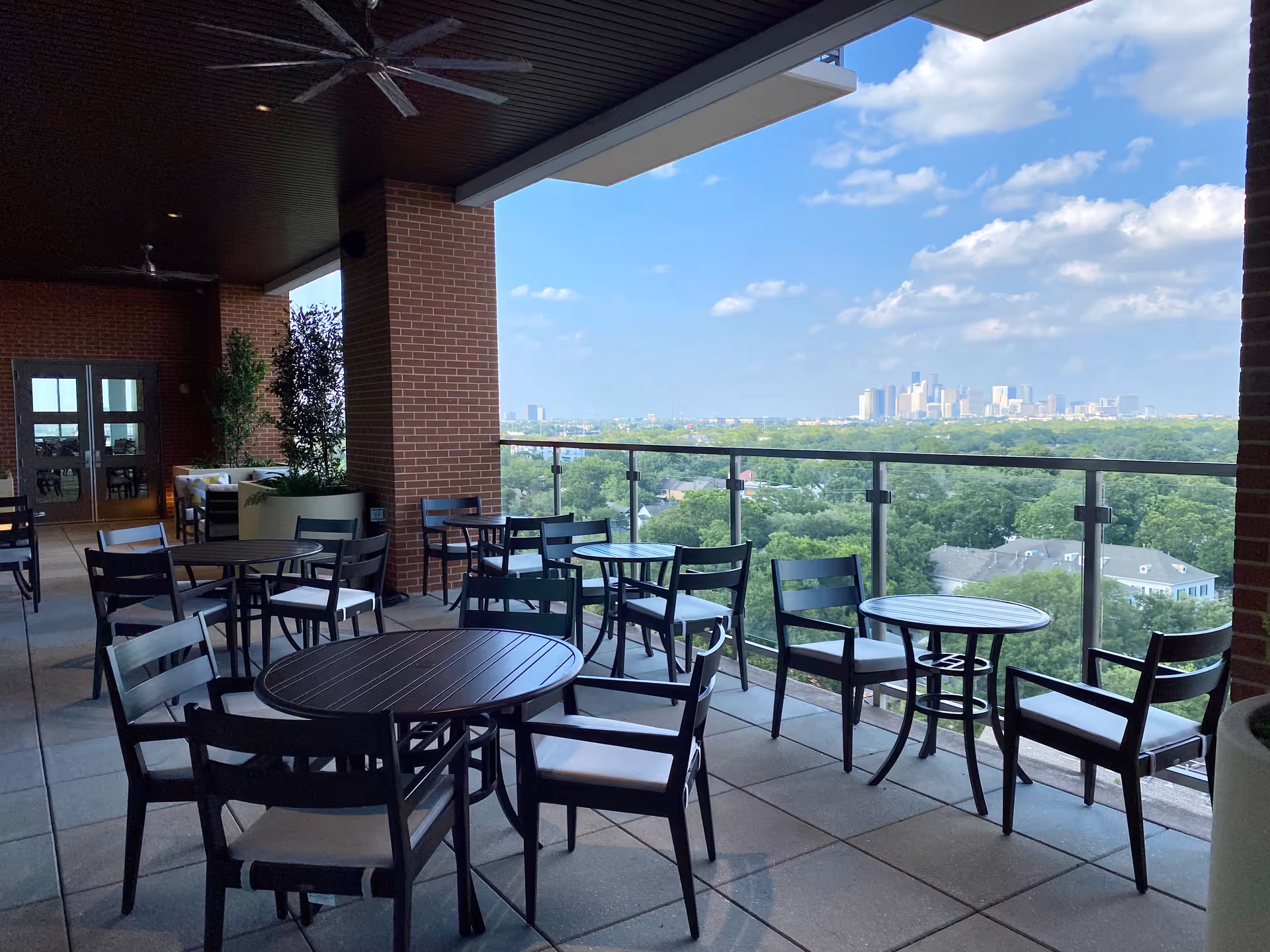 Outdoor covered patio area with several round tables and black chairs with white cushions. The patio has a glass railing overlooking a green landscape and a city skyline in the distance under a blue sky with scattered clouds. There are brick pillars and ceiling fans above the seating area.