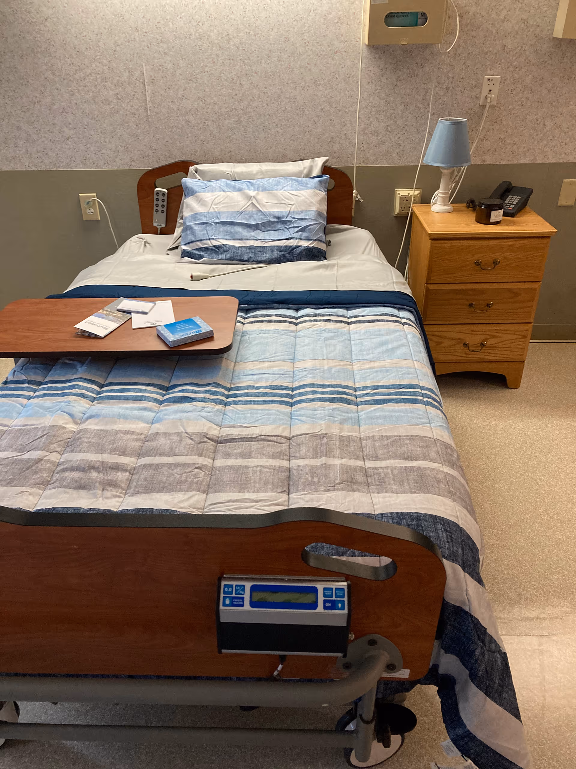 A hospital-style bed with a blue and gray striped comforter and matching pillows. There is a wooden bedside table with a blue lamp, a phone, and a small container. A wooden overbed table with some pamphlets and a box is placed on the bed. The room has a beige and gray wall with medical equipment mounted on it.