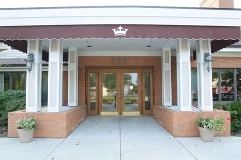 Entrance of Kingston Residence of Marion with double wooden doors under a maroon awning featuring a white crown symbol. The entrance is flanked by brick planters with green plants and flowers, and two lantern-style wall lights.