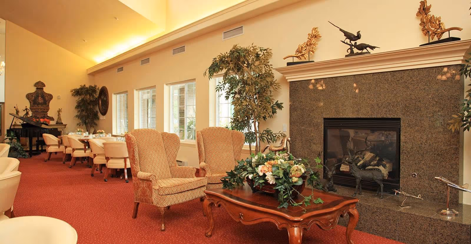 A cozy and elegant living room area with two patterned armchairs and a wooden coffee table adorned with a floral arrangement. The room features a large fireplace with decorative sculptures on the mantel, tall windows letting in natural light, and a red carpeted floor. In the background, there are several dining tables with cream-colored chairs and a grand piano near the wall.