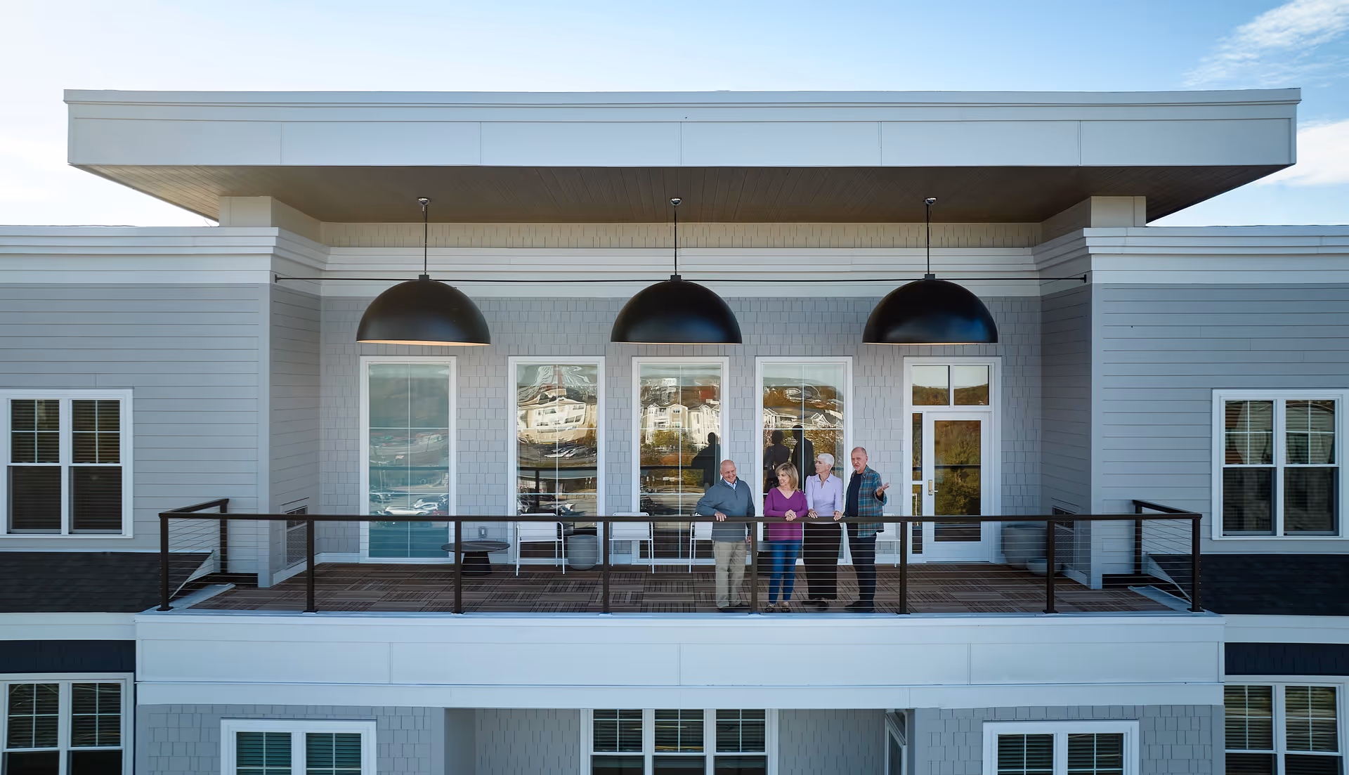 Four residents stand on a second-floor balcony of a modern senior living building beneath three large dome pendant lights.