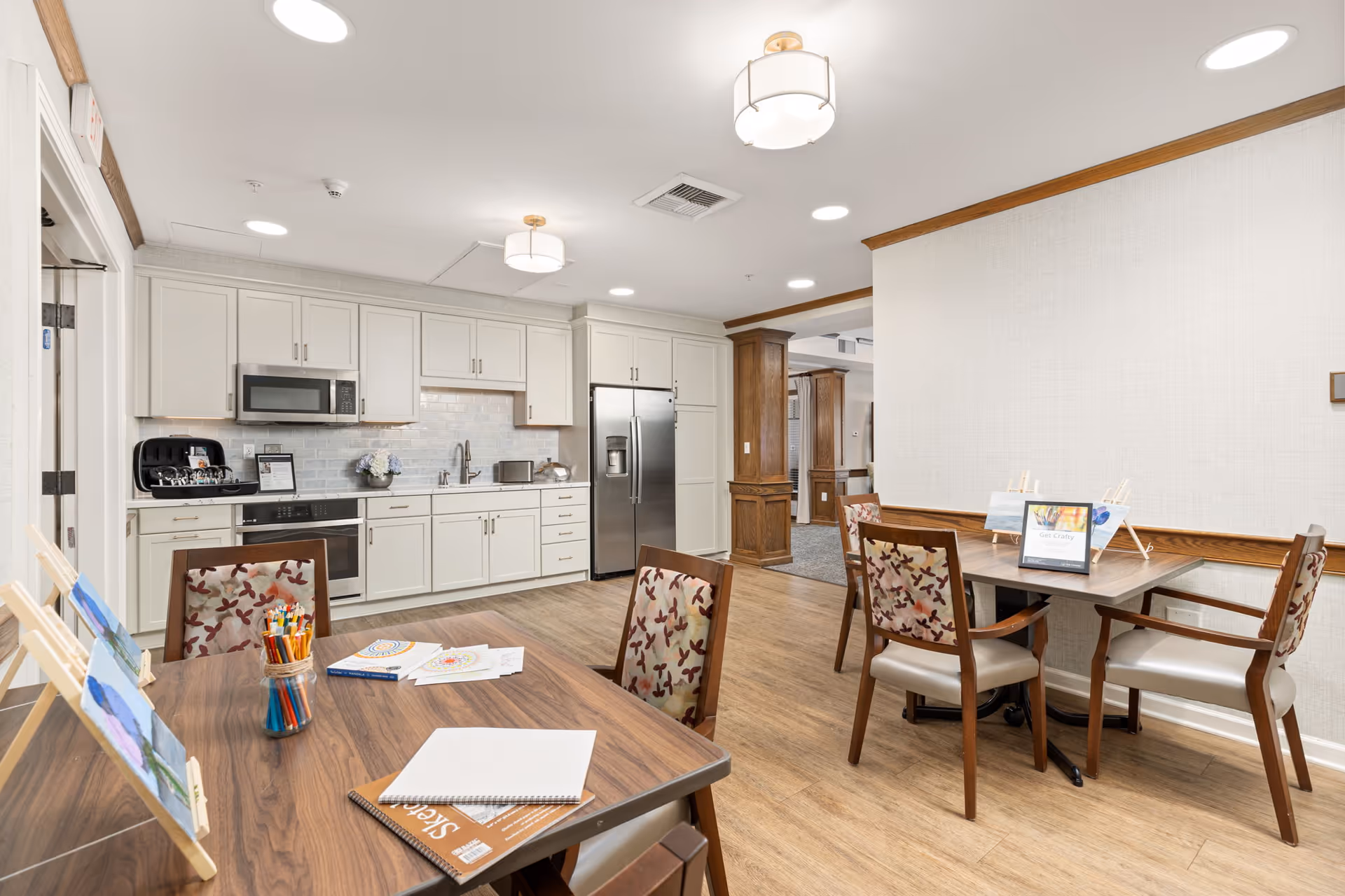 A bright and clean activity room with a kitchen area in the background. The kitchen features white cabinets, a stainless steel refrigerator, microwave, and oven. In the foreground, there are two wooden tables with chairs that have floral patterned upholstery. Art supplies, coloring books, and small easels with paintings are arranged on the tables, indicating a crafting or painting activity setup.