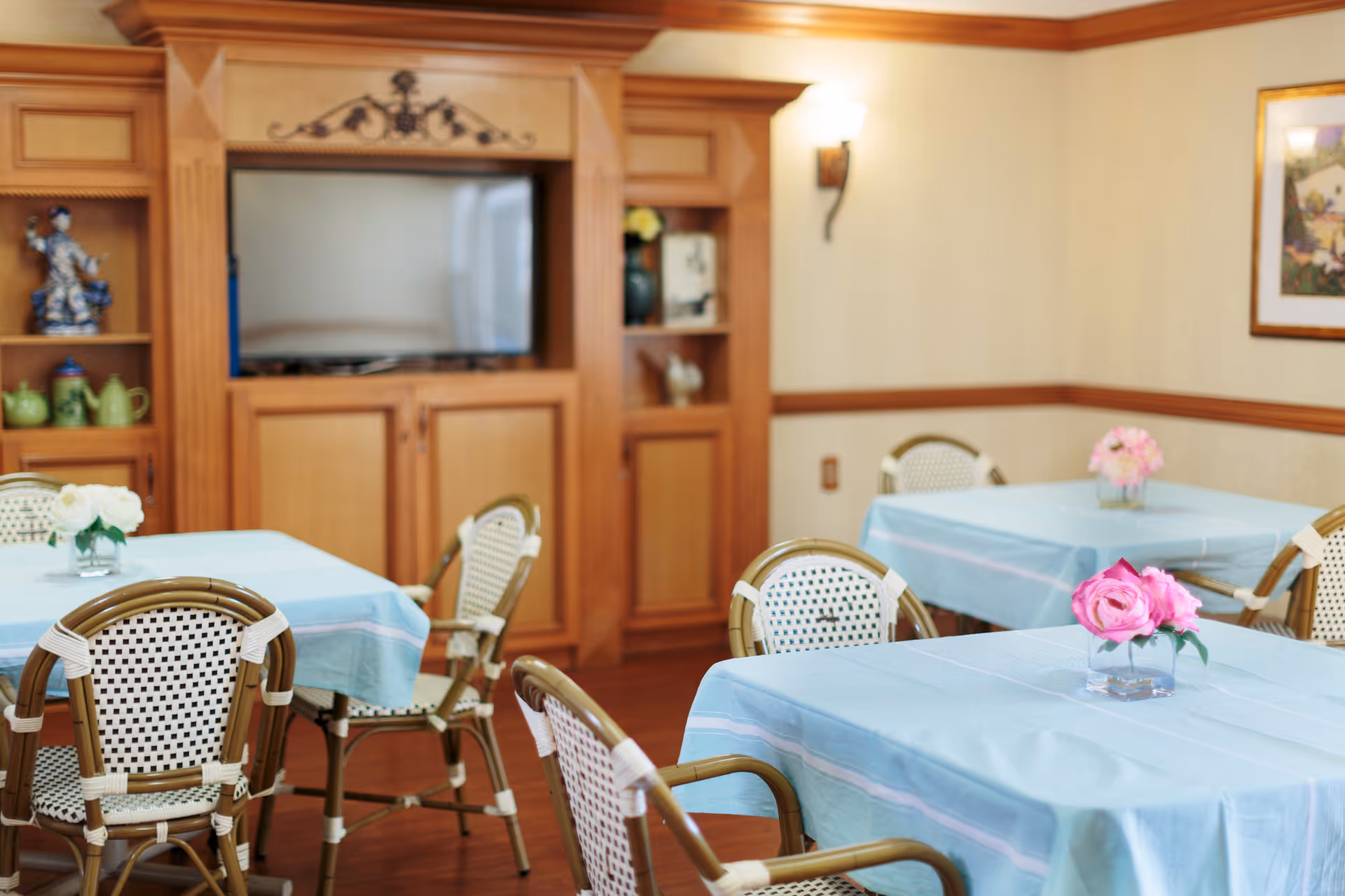 A dining room with tables covered in light blue tablecloths, each table decorated with a small vase of pink or white flowers. The room features wooden flooring, a wooden entertainment center with a TV, and light-colored walls with wood trim. Wicker chairs with white and brown woven seats and backs surround the tables.
