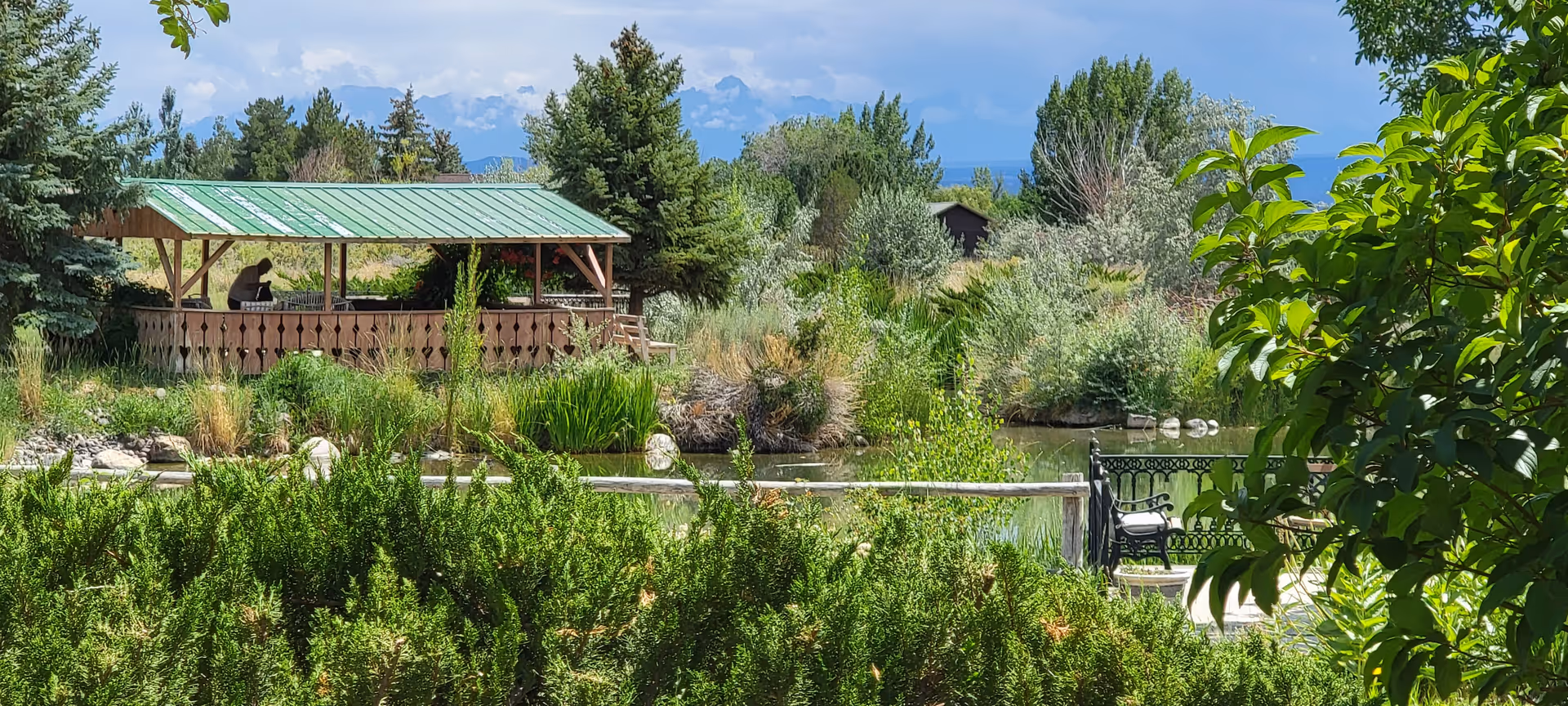 A wooden gazebo with a green metal roof beside a small pond, surrounded by shrubs, trees, a bench, and distant mountains.