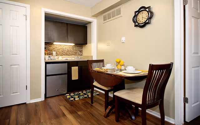 Small dining area with a two-seat table set beside a compact kitchenette with dark cabinets and wood flooring.