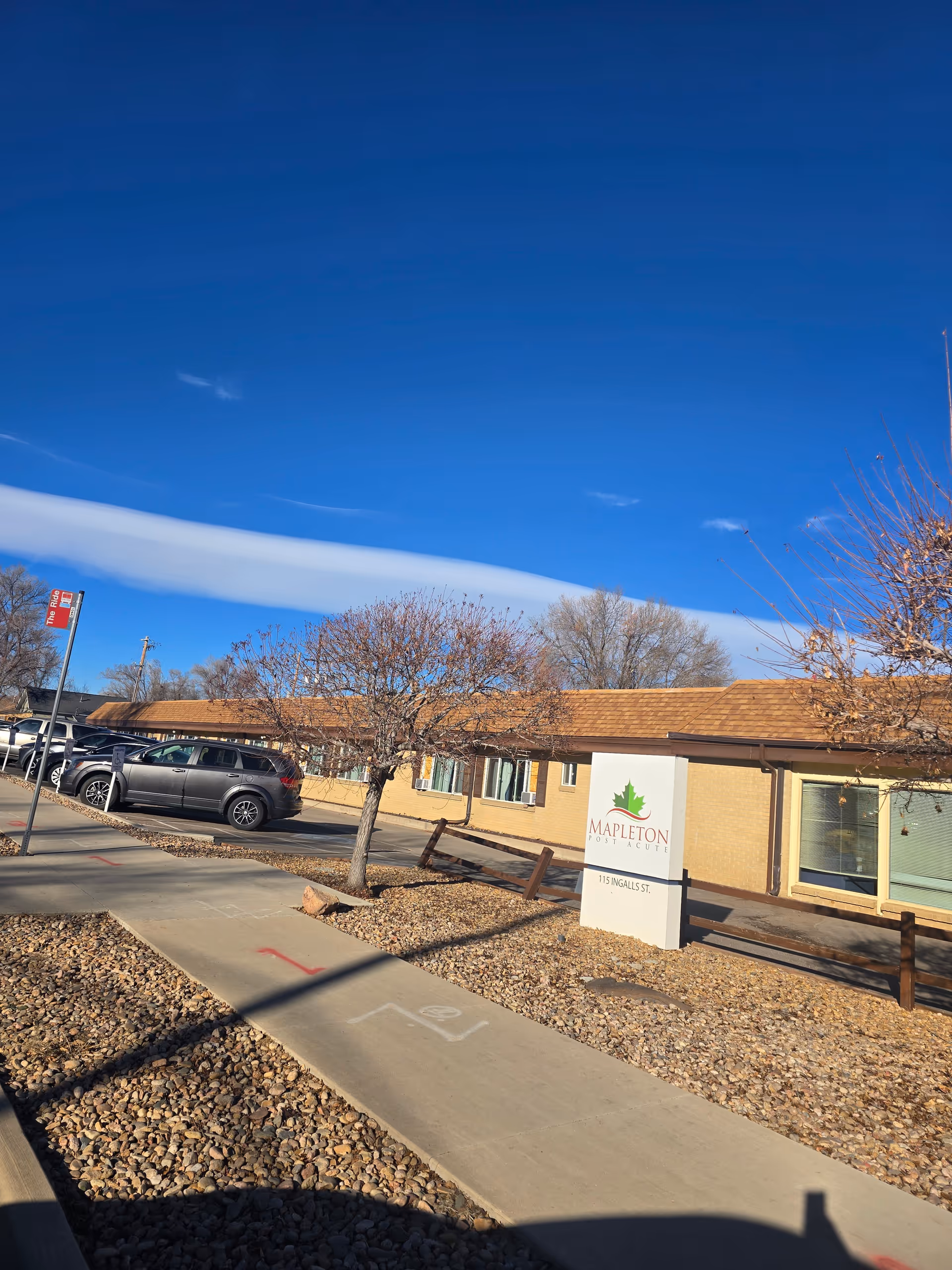 Exterior view of Mapleton Care Center building with a clear blue sky. There is a sidewalk and a parking area with several cars parked. Leafless trees and a sign with the facility's name and address are visible in front of the building.