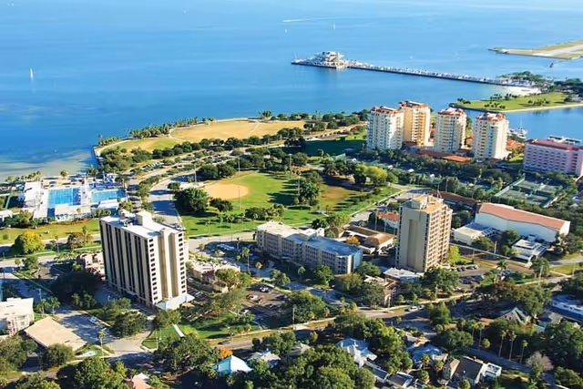 Aerial view of a coastal community featuring multiple mid-rise buildings, a baseball field, green spaces, and a pier extending into the water under a clear blue sky.
