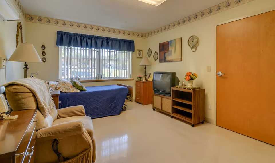 A cozy bedroom in a senior living facility with a single bed covered in a blue quilt, a beige recliner chair, a wooden dresser, and a TV on a wooden stand. The room has a large window with vertical blinds and a blue valance, floral wallpaper border near the ceiling, and various decorative items including a wall clock and framed pictures.