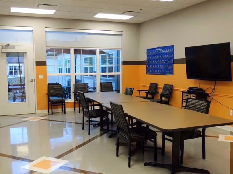 A meeting or activity room with a long rectangular table surrounded by black chairs. The walls are painted yellow with a brown stripe, and there is a large window and a glass door letting in natural light. A flat-screen TV is mounted on the wall, and a blue chart or poster is displayed next to it.