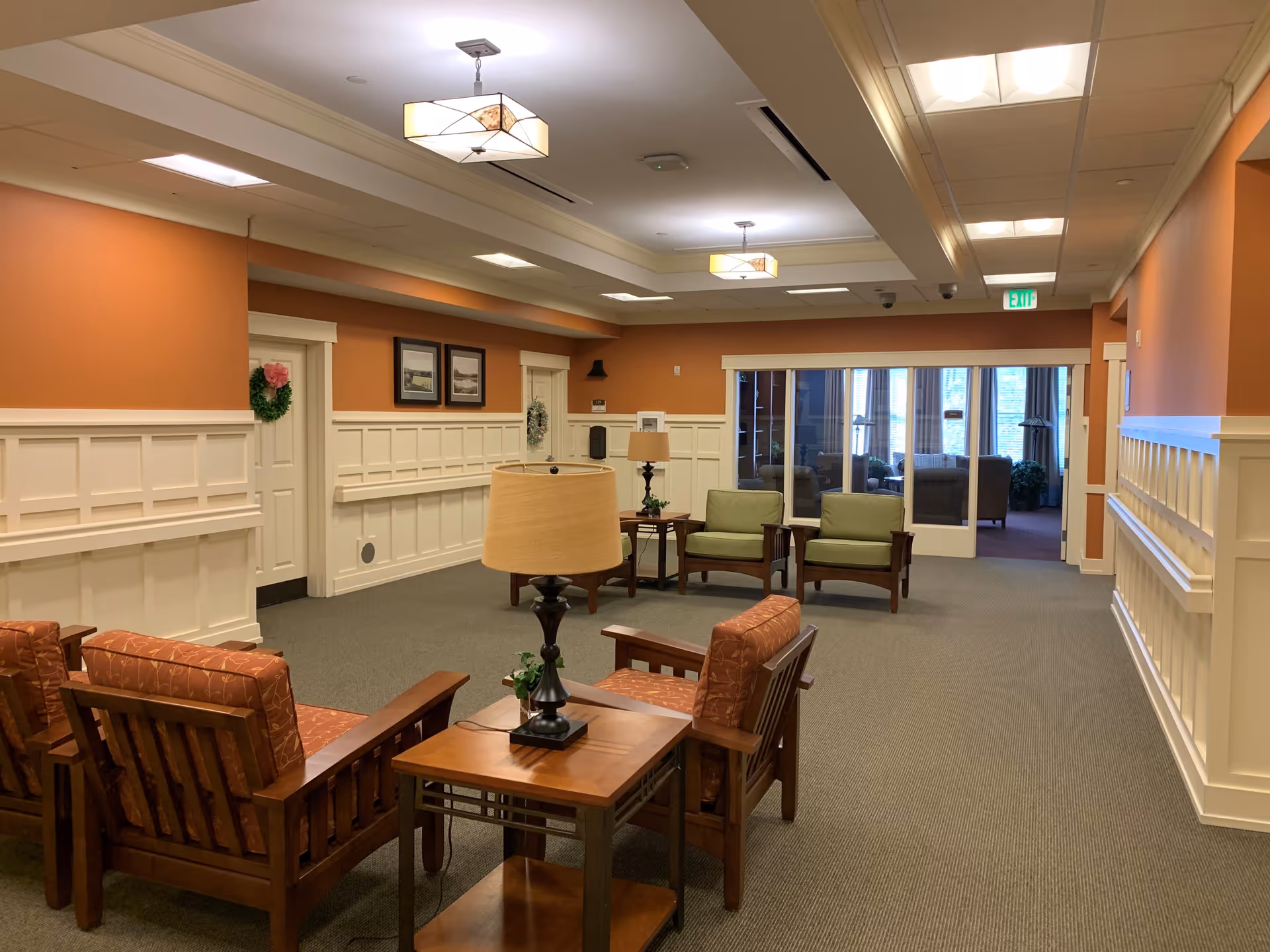 A cozy interior common area with orange walls and white wainscoting. The room features wooden chairs with cushions in orange and green, small wooden tables with lamps, framed pictures on the wall, and a glass door leading to another seating area with large windows.