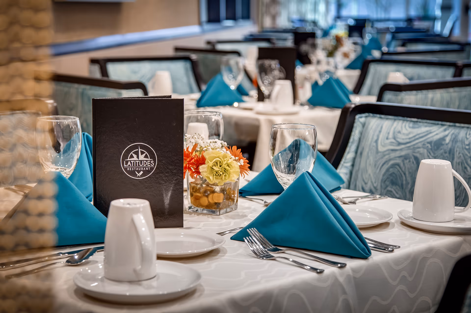 A neatly set dining table in a restaurant with white tablecloth, teal folded napkins, clear wine glasses, white coffee mugs on saucers, silverware, a small floral centerpiece with orange and yellow flowers, and a black menu card with the logo 'Latitudes Restaurant'. Upholstered chairs with a blue and white pattern surround the tables.
