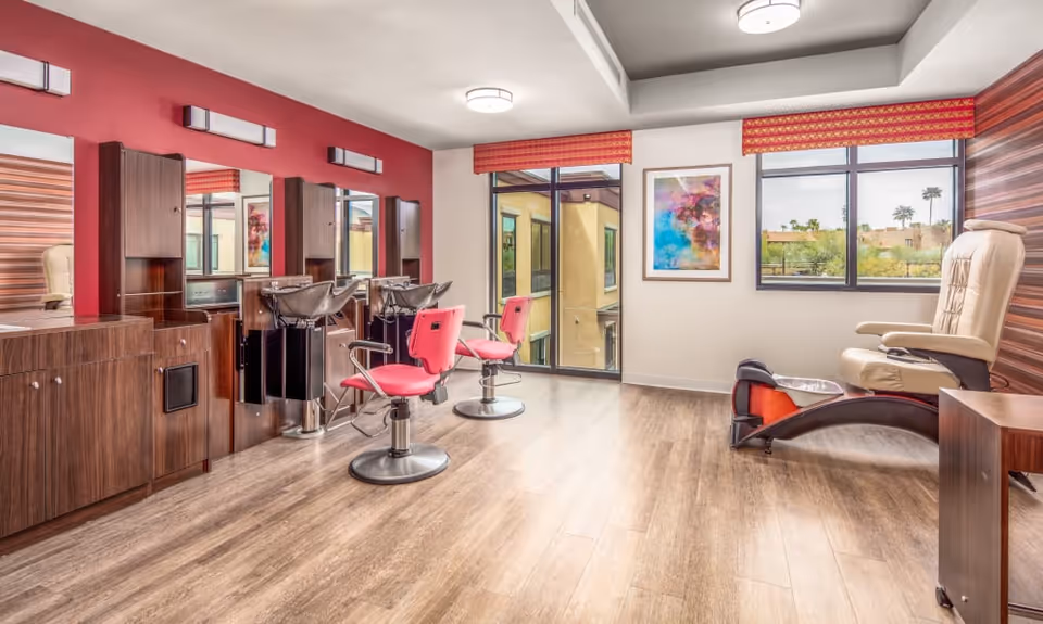 Interior view of a salon room in a senior living facility with two pink salon chairs in front of mirrors and washbasins, a beige pedicure chair with a foot bath, wooden cabinets, large windows with red valances, and a colorful abstract painting on the wall.
