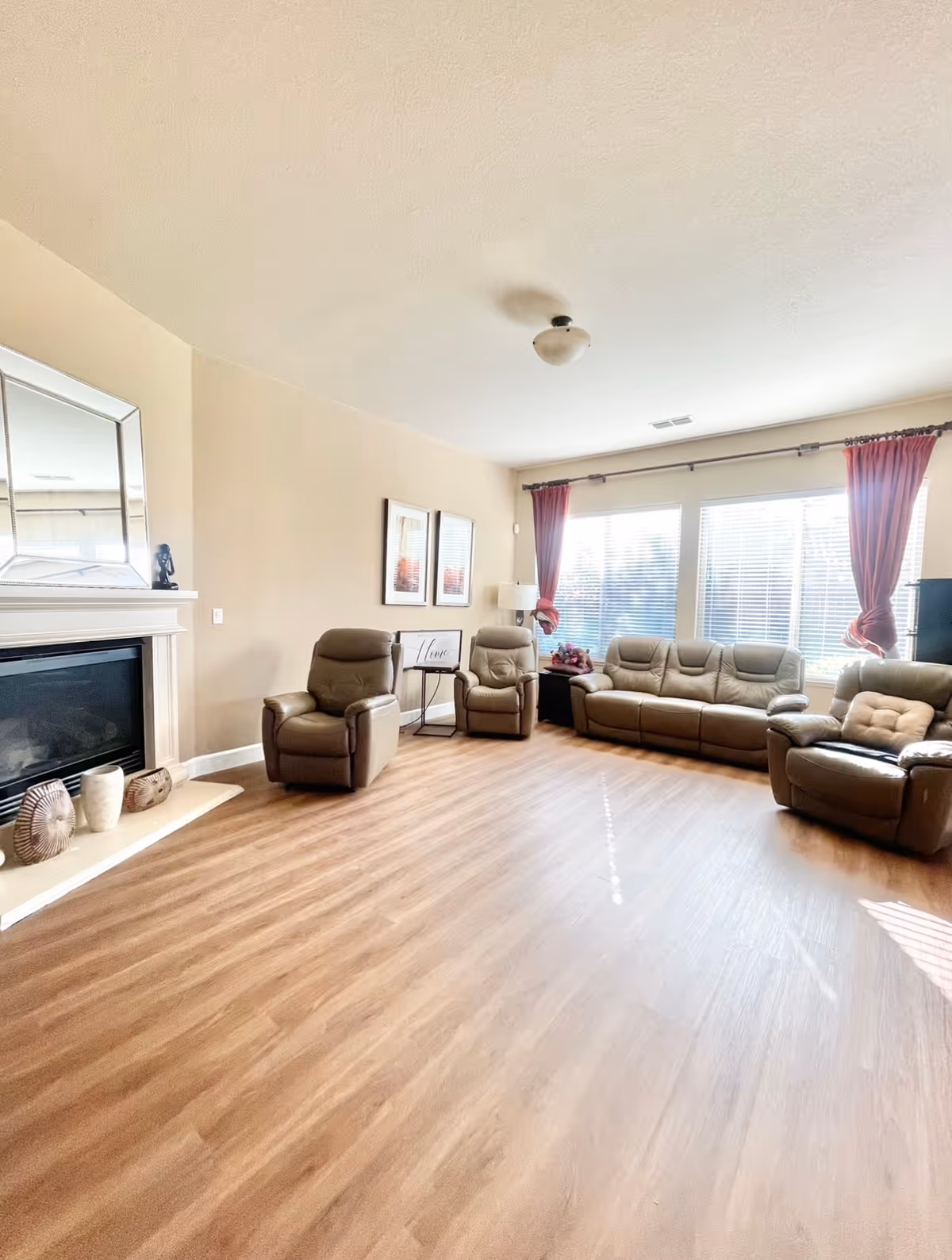 A bright and spacious living room with light wood flooring, beige walls, and a white ceiling. The room features a fireplace with decorative items on the hearth, a large mirror above it, and three brown leather recliners arranged around the room. There is a brown leather sofa near large windows with white blinds and red curtains, allowing natural light to fill the space. Two framed pictures and a small table with a lamp and decorative sign are on the wall adjacent to the sofa.