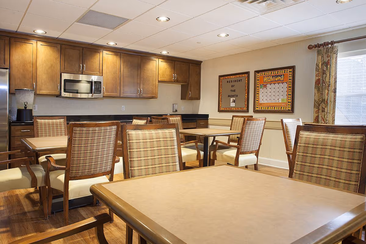 A well-lit dining room with several tables and cushioned chairs featuring plaid upholstery. The room has wooden cabinetry along one wall with a microwave and a refrigerator. Two framed bulletin boards hang on the wall, one labeled 'Resident of the Month' and the other displaying a calendar. A window with floral curtains allows natural light into the room.