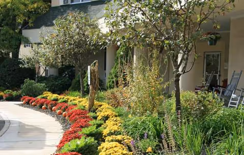 Colorful flower beds line a curved walkway beside a building with a small porch.