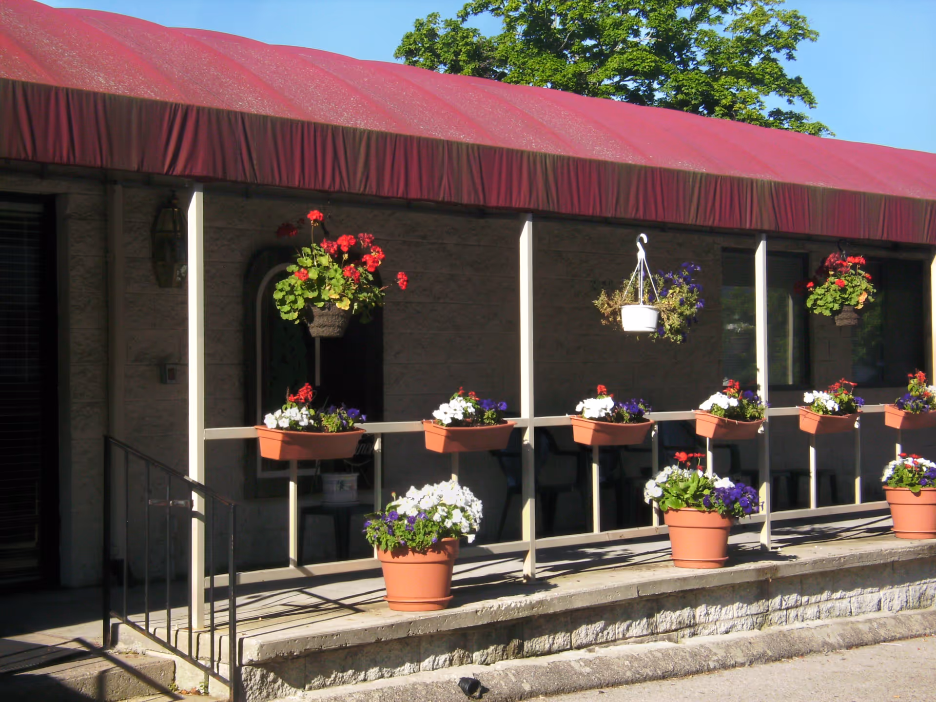 Outdoor view of a building entrance with a red awning and a railing decorated with multiple flower pots containing colorful flowers. There are also hanging flower baskets and some greenery visible in the background.