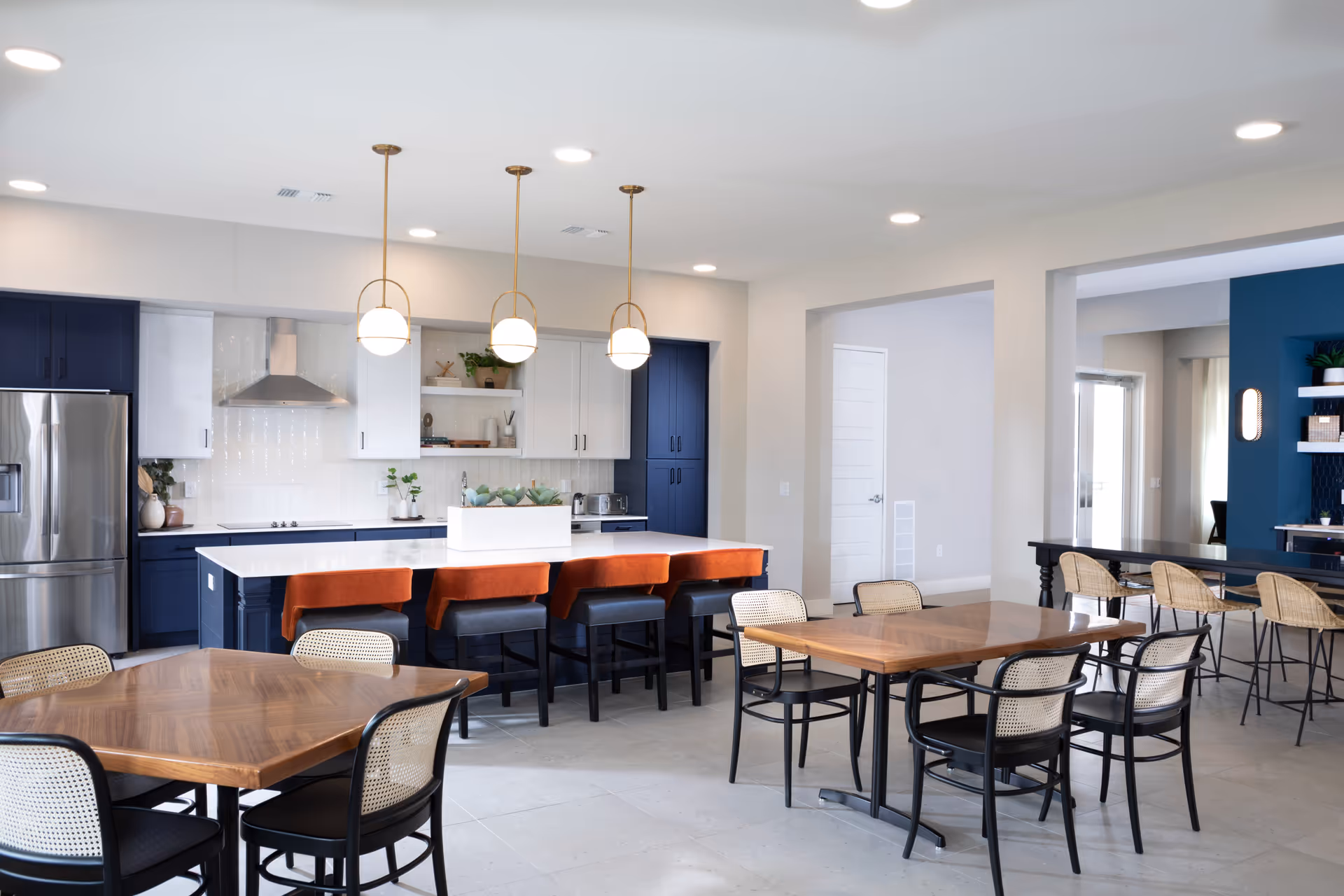 A modern communal dining and kitchen area with wooden tables and black chairs. The kitchen features navy blue lower cabinets, white upper cabinets, a stainless steel refrigerator, and a range hood. There is a large island with a white countertop and four orange cushioned bar stools. Three pendant lights hang above the island. The room has a clean, bright, and open design with light-colored walls and tiled flooring.