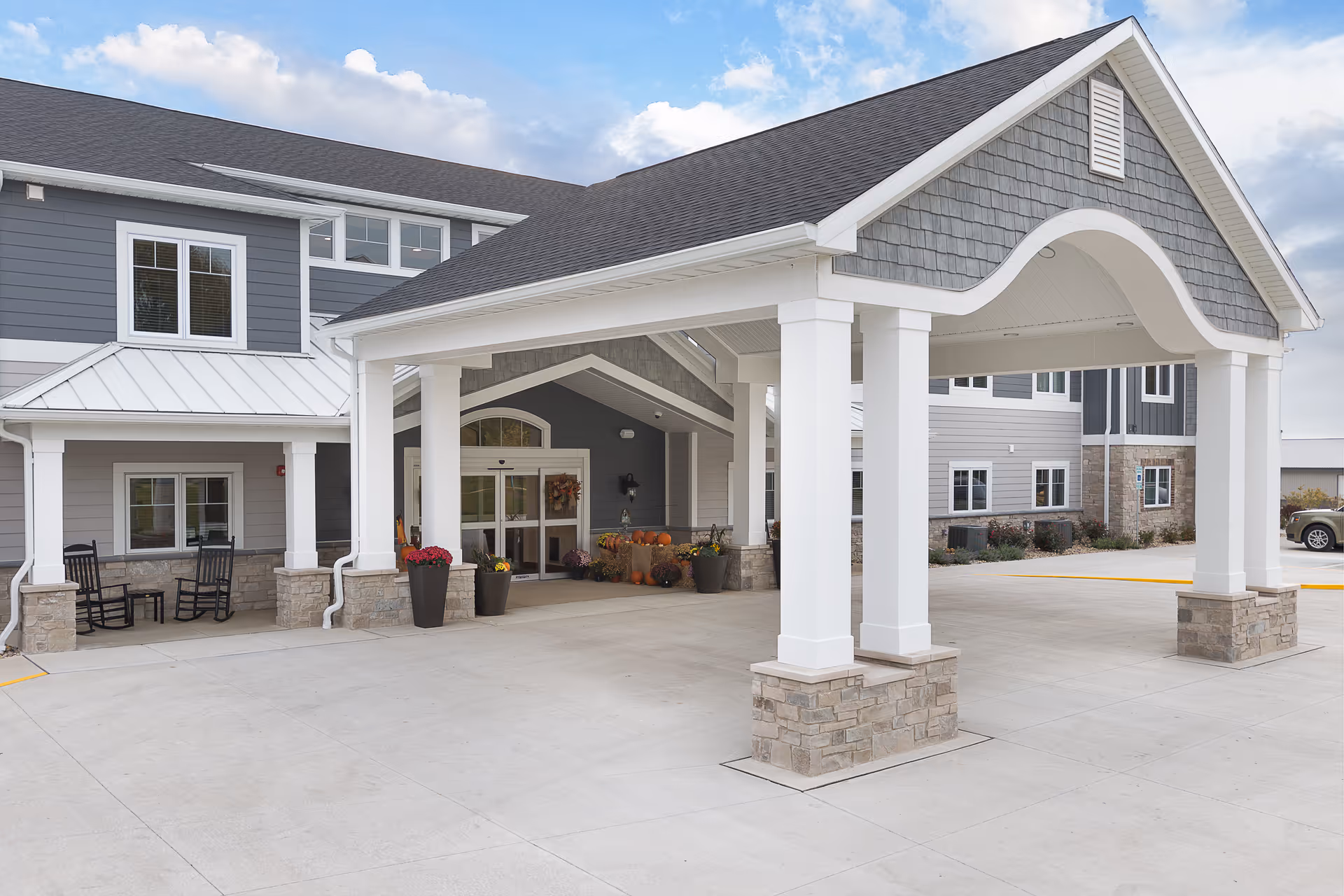 Entrance of a senior living facility with a covered drop-off area supported by white columns with stone bases. The building exterior features gray siding, white trim, and multiple windows. There are rocking chairs on the porch and potted plants with flowers and pumpkins near the entrance door under a partly cloudy sky.