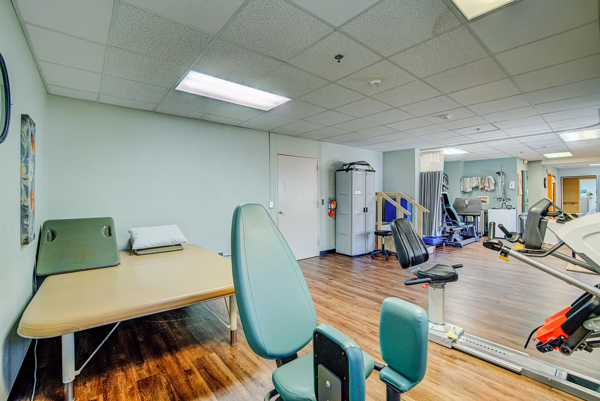 A rehabilitation or physical therapy room with exercise equipment, a padded treatment table with a pillow, chairs, and medical supplies. The room has wood flooring, light green walls, and a drop ceiling with fluorescent lighting.