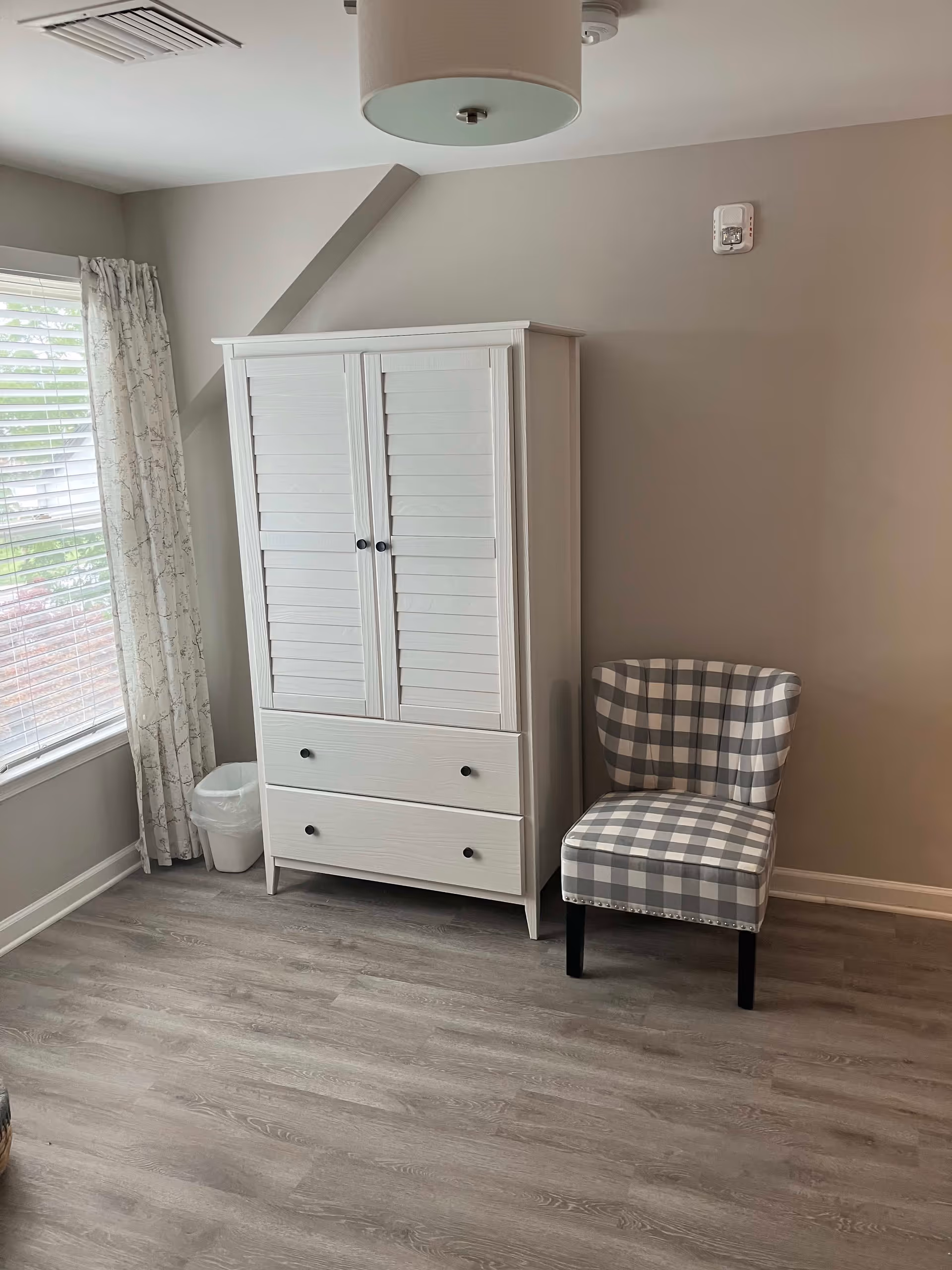 A corner of a room with a white wooden wardrobe with two drawers and two doors, a gray and white checkered upholstered chair, a window with white blinds and patterned curtains, and light wood flooring.