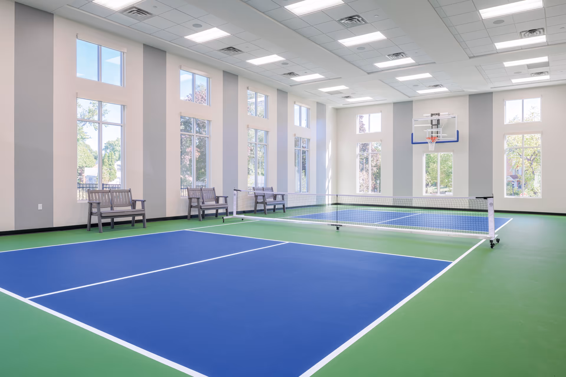 Indoor sports court with a blue and green floor marked for pickleball, a net in the center, and a basketball hoop mounted on the far wall. The room has tall windows letting in natural light and several benches along the walls.