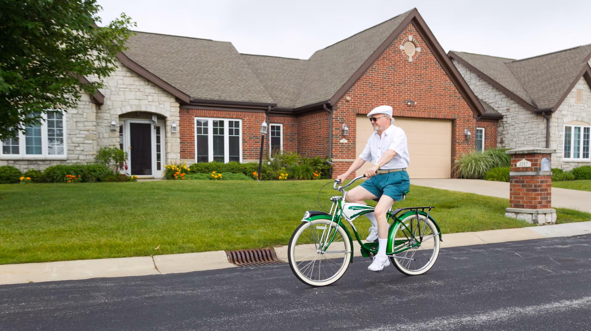 A man rides a green bicycle along a street in front of a single-story brick and stone house with a garage and lawn.