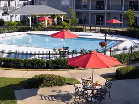 Outdoor swimming pool area with several red umbrellas and black metal tables and chairs. The pool is surrounded by a black metal fence and greenery. In the background, there is a covered pavilion and a multi-story building with balconies.