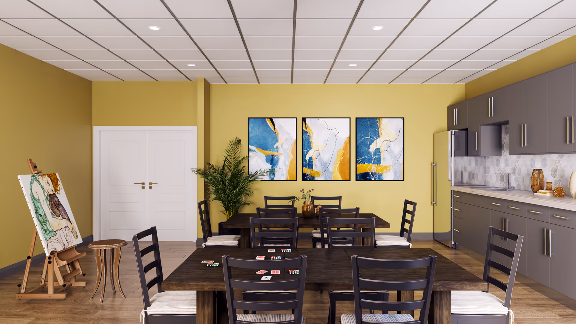 A bright, modern common room with yellow walls and a tiled ceiling. The room features two dark wooden tables with several chairs around them, some with playing cards and poker chips on the table. On the right side, there is a kitchen area with gray cabinets, a refrigerator, and a countertop with decorative items. On the left side, there is an easel with a painting and a small wooden stool. Three abstract paintings in blue, white, and gold tones hang on the wall above a potted plant.