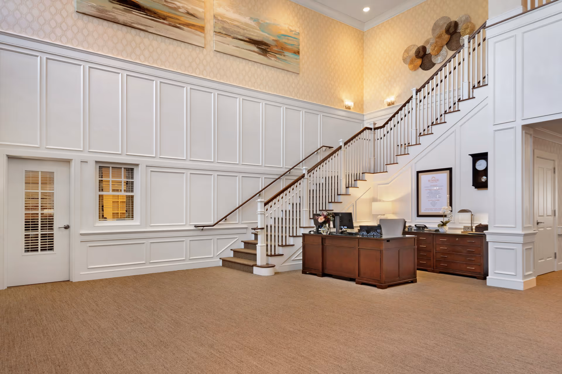 Interior view of a senior living facility reception area with a wooden front desk, a staircase with white railings, beige carpet, and decorative wall art above the staircase.