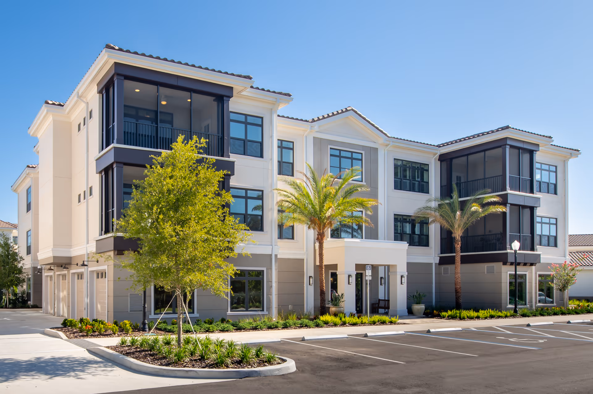 Exterior view of a modern three-story senior living facility building with large windows, balconies, palm trees, and a clear blue sky. The building has a beige and gray facade with a tiled roof and a parking area in front.