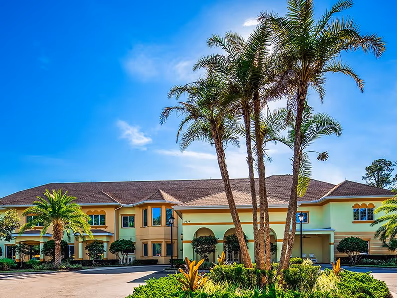 Exterior view of The Windsor at Ortega senior living facility with a clear blue sky, palm trees, and well-maintained landscaping in front of a two-story building with a brown roof and light yellow walls.