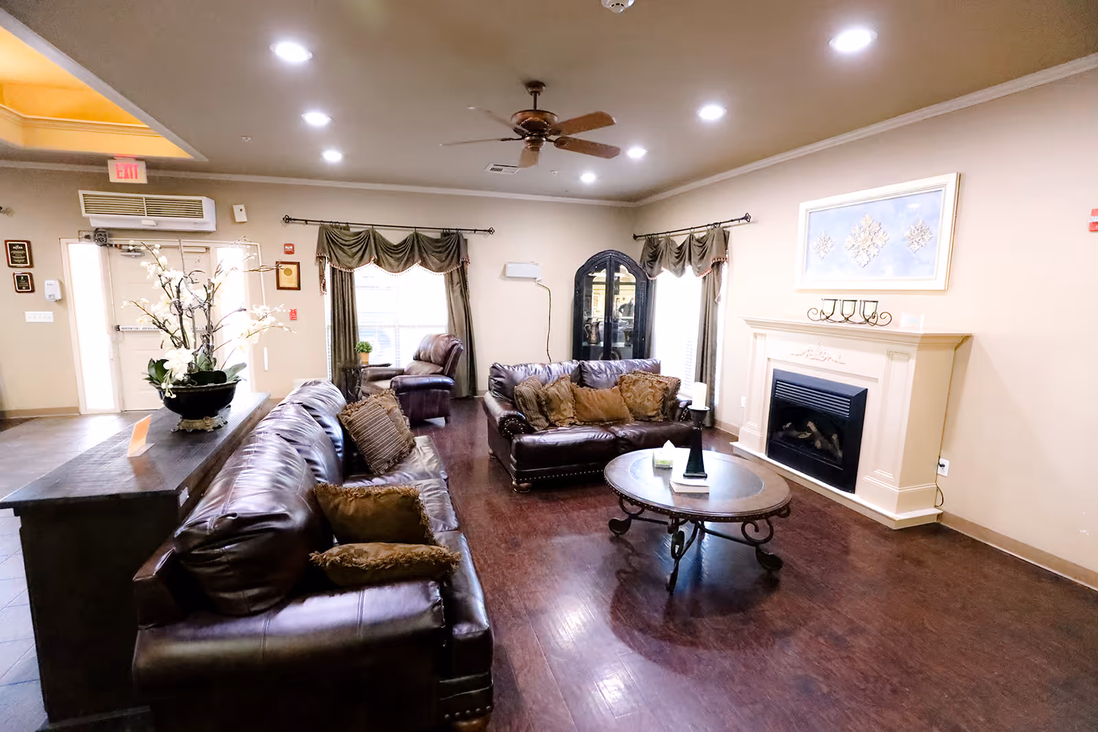 A cozy living room area with two brown leather sofas, a round wooden coffee table, and a fireplace with a decorative mantle. The room has hardwood floors, a ceiling fan, and windows with green curtains. There is a dark wooden cabinet in the corner and a flower arrangement on a side table near the entrance.