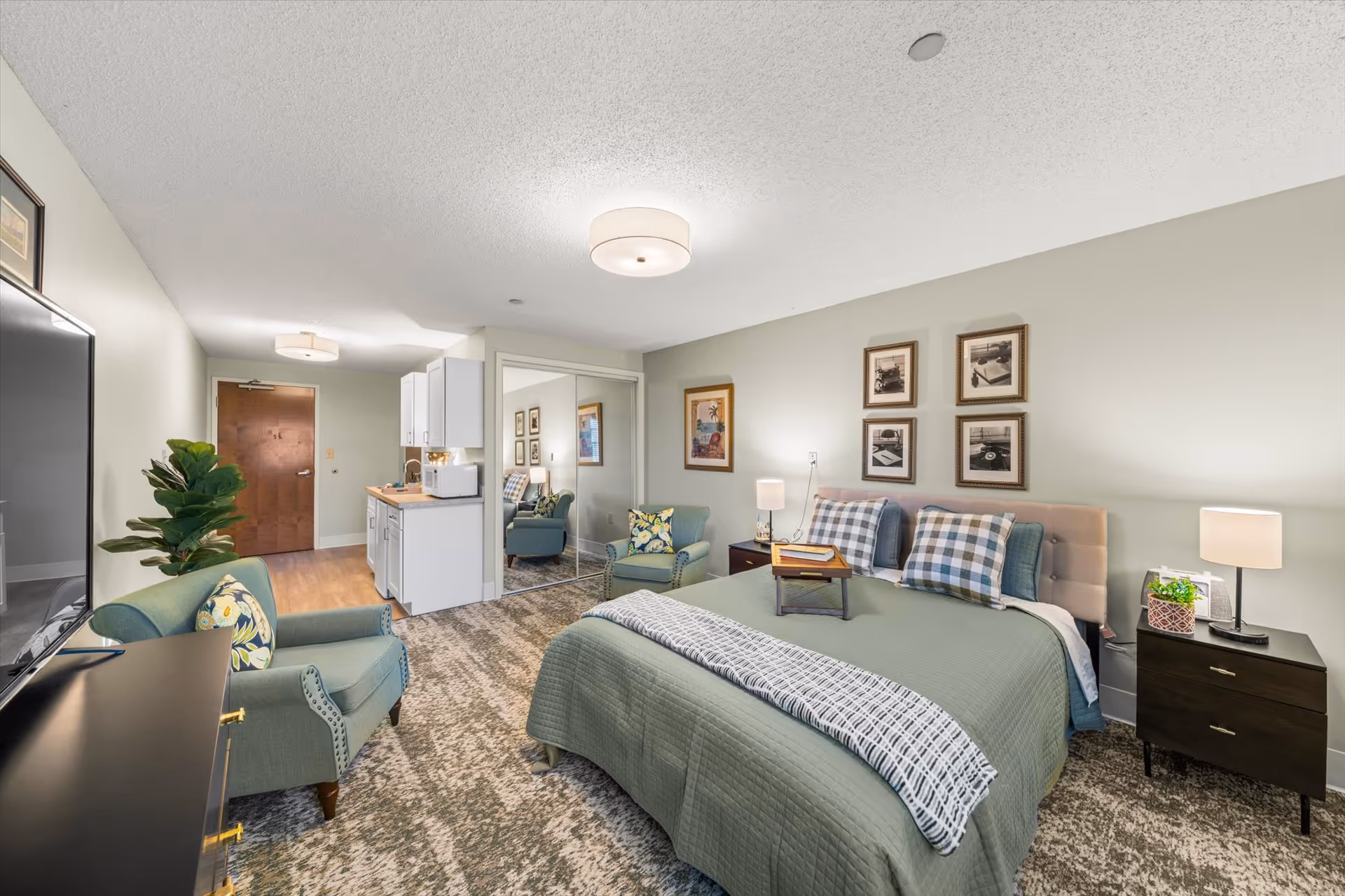 A cozy bedroom in a senior living facility with a large bed covered in green bedding and plaid pillows. The room features a comfortable armchair with a floral cushion, a flat-screen TV on a black dresser, a small kitchenette with white cabinets, and a mirrored closet door. The walls are decorated with framed pictures, and there are two bedside tables with lamps. The floor is carpeted with a patterned design.